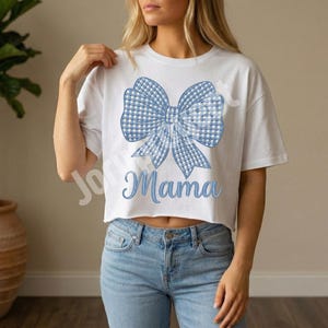 Puede incluir: Top corto blanco con un gran lazo de cuadros azul y la palabra "Mama". El lazo tiene un patrón de cuadros azul claro y blanco. La camiseta se combina con vaqueros azules.