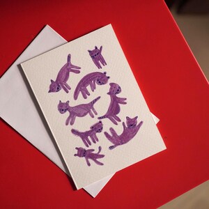 May include: A greeting card with a white envelope. The card features a whimsical illustration of purple cats in various poses. The card and envelope are on a red surface.