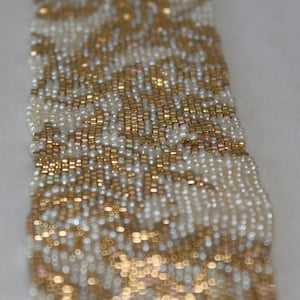 Puede incluir: Un brazalete de cuentas doradas y blancas con un patrón floral. El brazalete está hecho de pequeñas cuentas redondas que se tejen juntas para crear un diseño delicado e intrincado.