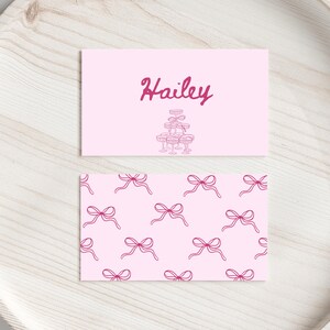 Op de afbeelding: Twee roze kaarten op een lichtroze houten dienblad. Eén kaart heeft de naam "Hailey" in cursief met een champagne toren illustratie. De andere kaart heeft een patroon van roze strikken. De kaarten zijn waarschijnlijk voor een feest of evenement.