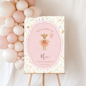 May include: A welcome sign for a first birthday celebration. The sign features a cartoon mouse in a tutu holding a heart-shaped balloon. The sign is pink and cream-colored with the text "Welcome to Mia's First Birthday 22.09.2025". A balloon arch is on the left.