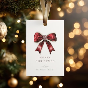 Op de afbeelding: Een witte kerstcadeaulabel met een aquarel illustratie van een rode strik met glitter en witte rand. Het label heeft de woorden "MERRY CHRISTMAS" en "with Love" en een achternaam. Een gouden lint is bevestigd.
