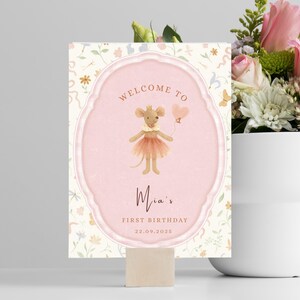 May include: A decorative sign with a pink oval frame and a watercolor illustration of a mouse holding a heart-shaped balloon. The sign reads "Welcome to Mia's First Birthday 22.09.2025". The background has a floral pattern.