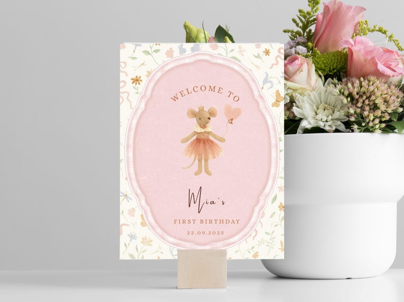 May include: A decorative sign with a pink oval frame and a watercolor illustration of a mouse holding a heart-shaped balloon. The sign reads "Welcome to Mia's First Birthday 22.09.2025". The background has a floral pattern.