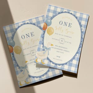 Może przedstawiać: Dwa zaproszenia na pierwsze urodziny. Zaproszenia mają niebieski wz&oacute;r w kratkę i owalny kształt z napisem "ONE Silly Goose". Balony i ilustracja gęsi są r&oacute;wnież dołączone.