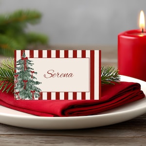 Peut inclure: Une carte de place de Noël avec un motif d'arbre de Noël à l'aquarelle et le nom "Serena". La carte est posée sur une serviette rouge, avec une bougie rouge allumée et une branche de pin. Le fond est une table en bois.
