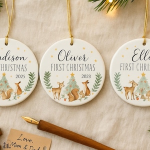 Op de afbeelding: Drie witte keramische kerstversieringen met gouden ophangkoorden. Elk ornament heeft een naam, de tekst "FIRST CHRISTMAS" en het jaar. De ornamenten hebben illustraties van bosdieren en kerstbomen.