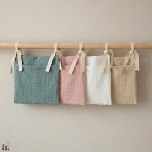 May include: Four fabric storage pockets in shades of teal, pink, white, and beige hang from a wooden rod. Each pocket is rectangular with a front pocket and is tied to the rod with cream-colored ribbons. The pockets are made of a textured, gauzy fabric.