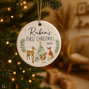 Op de afbeelding: Een ronde, witte kerstbal met de tekst "Ruben's First Christmas 2025". De ornamenten hebben een illustratie van een hert, vos, eekhoorn en kerstboom. Een gouden touwtje is bevestigd om op te hangen.