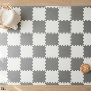 Könnte beinhalten: Eine graue und weiße, ineinandergreifende Spielmatte aus Schaumstoff mit Schachbrettmuster. Ein Weidenkorb mit einer cremefarbenen Decke und ein Holzspielzeug befinden sich in den oberen Ecken. Die Matte liegt auf einem hellen Holzboden.