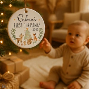 Op de afbeelding: Een ronde, witte kerstversiering met de tekst "Ruben's FIRST CHRISTMAS 2025". De versiering toont een aquarel illustratie van een hert, vos, eekhoorn en kerstboom. Een gouden touwtje is bevestigd om op te hangen.