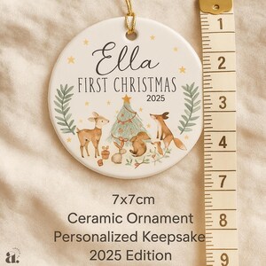 Op de afbeelding: Een witte keramische kerstversiering met een gouden touwtje, met de tekst "Ella FIRST CHRISTMAS 2025". De versiering heeft een boslandschap met een hert, een vos en een kerstboom. Het meet 7 x 7 cm.