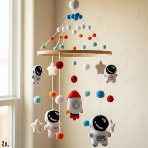 May include: A nursery mobile with a wooden ring, felt astronauts, stars, a rocket, and colourful pom-poms. The mobile is suspended by white strings and features blue, orange, red, and white accents.
