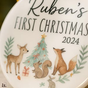 Op de afbeelding: Een witte keramische kerstversiering met de tekst "Ruben's FIRST CHRISTMAS 2024". De versiering heeft een aquarel illustratie van een hert, vos, eekhoorn, konijn, kerstboom en cadeau.
