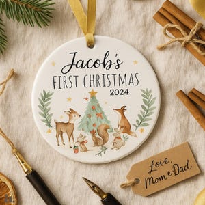 Op de afbeelding: Een witte keramische kerstbal met de tekst "Jacob's FIRST CHRISTMAS 2024". De bal heeft een aquarel illustratie van bosdieren rond een kerstboom. Een gouden lint is bevestigd om op te hangen.