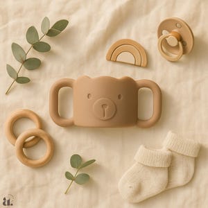 May include: A selection of baby essentials in beige and brown tones. Features a bear-faced sippy cup, a dummy, a wooden rainbow, two wooden rings, and a pair of cream socks. Eucalyptus sprigs are arranged around the items.