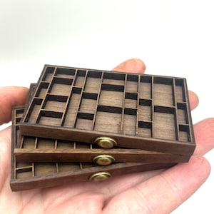 Peut inclure: Trois plateaux rectangulaires en bois empilés avec une finition brun foncé et de multiples petits compartiments. Chaque plateau présente un bouton central de couleur laiton sur le bord avant. Les plateaux sont dotés de séparateurs.