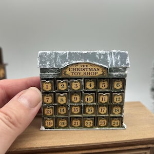 Könnte beinhalten: Ein Miniatur-Adventskalender in Form eines Gebäudes mit grauem Dach und vierundzwanzig kleinen Schubladen, die von eins bis vierundzwanzig nummeriert sind. Das Schild über den Schubladen trägt die Aufschrift THE CHRISTMAS TOY SHOP in Schwarz.