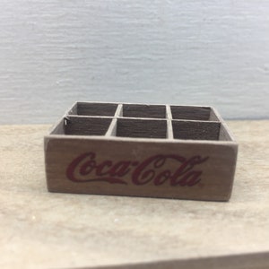 May include: A miniature wooden crate with six compartments, painted brown and featuring the Coca-Cola logo in red.