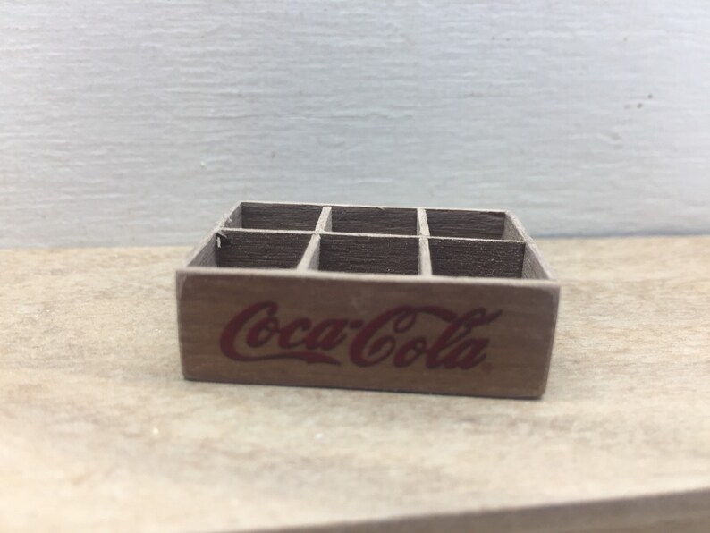 May include: A miniature wooden crate with six compartments, painted brown and featuring the Coca-Cola logo in red.