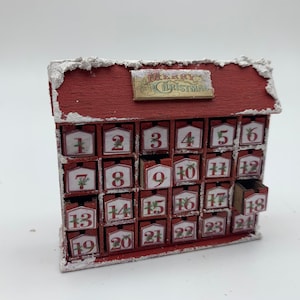 May include: A miniature red wooden Christmas advent calendar with 24 numbered drawers. The roof is trimmed with white, and a small sign reads "Merry Christmas". One drawer is open, revealing its interior.
