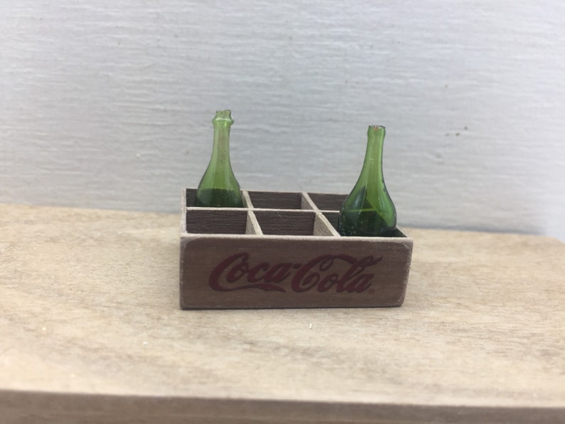 May include: A miniature wooden Coca-Cola crate with two green glass bottles inside.
