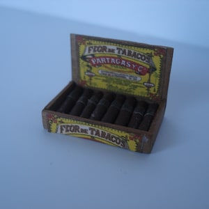 May include: A wooden box of cigars with a yellow and red label that reads "FLOR DE TABACOS PARTAGASY C".