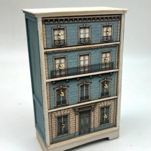 Könnte beinhalten: Eine kleine Holzkommode mit einer aufgedruckten Fassade eines vierstöckigen Gebäudes. Die blau-beige Außenansicht zeigt Fenster, einen Balkon und eine zentrale Tür. Jede Schublade ist mit einem kleinen, runden, goldfarbenen Metallknopf versehen.