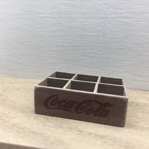 May include: A wooden crate with six compartments, designed to hold six bottles. The crate is painted brown and has the words "Coca-Cola" printed in red on the front.