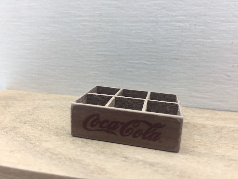 May include: A wooden crate with six compartments, designed to hold six bottles. The crate is painted brown and has the words "Coca-Cola" printed in red on the front.