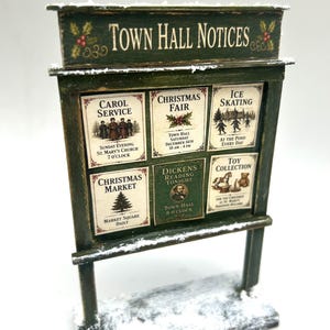 Può includere: Una bacheca in miniatura in legno verde con una finitura innevata. Il cartello riporta la scritta TOWN HALL NOTICES e mostra cinque poster individuali per eventi che includono un servizio di canti natalizi, una fiera e il pattinaggio su ghiaccio.