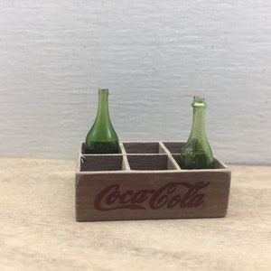 May include: A wooden Coca-Cola crate with two green glass bottles inside. The crate has a red Coca-Cola logo printed on the front.