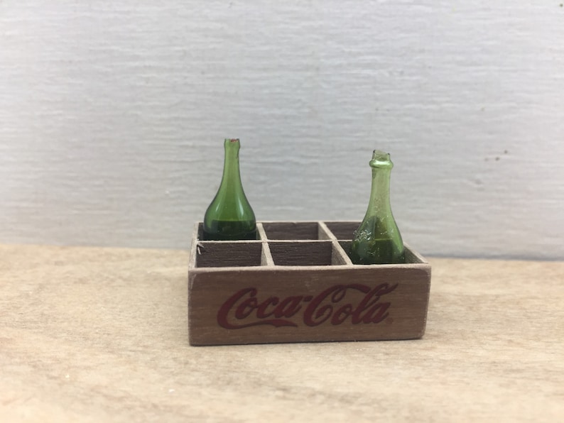 May include: A wooden Coca-Cola crate with two green glass bottles inside. The crate has a red Coca-Cola logo printed on the front.