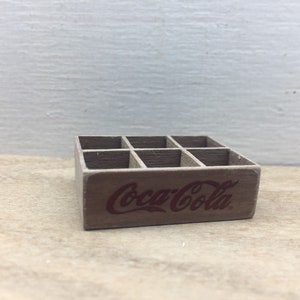 May include: A miniature wooden crate with six compartments, painted brown and featuring the Coca-Cola logo in red.