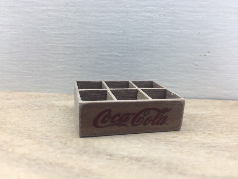 May include: A miniature wooden crate with six compartments, painted brown and featuring the Coca-Cola logo in red.