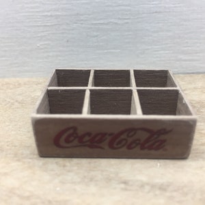 May include: A miniature wooden crate with six compartments, featuring the Coca-Cola logo in red lettering.