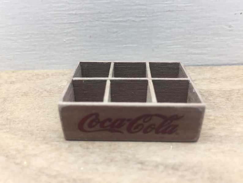 May include: A miniature wooden crate with six compartments, featuring the Coca-Cola logo in red lettering.