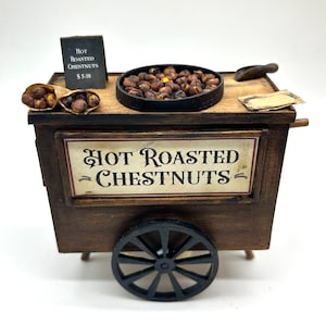 May include: A miniature wooden chestnut cart with a black metal pan filled with roasted chestnuts. The cart has a sign that reads "Hot Roasted Chestnuts" and a small chalkboard sign with the price. A paper cone of chestnuts sits on the cart.