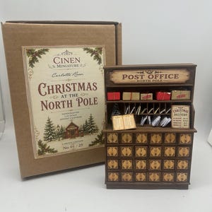 Puede incluir: Un calendario de adviento en miniatura de madera con forma de oficina de correos, con veinticuatro cajones numerados, estantes con paquetes pequeños, ranuras para correo y un letrero que dice Post Office North Pole, junto a su caja.