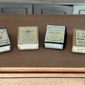 Może przedstawiać: Cztery miniaturowe, zabytkowe pudełka z akcesoriami do szycia, w tym guziki i nici. Pudełka są prostokątne z etykietami w języku francuskim i angielskim. Pudełka są w różnych kolorach, w tym czarnym, złotym i białym. Pudełka są ułożone na drewnianej powierzchni.