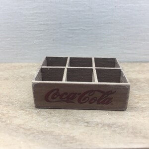 May include: A wooden Coca-Cola crate with six compartments. The crate is brown and has the Coca-Cola logo printed on the front in red.