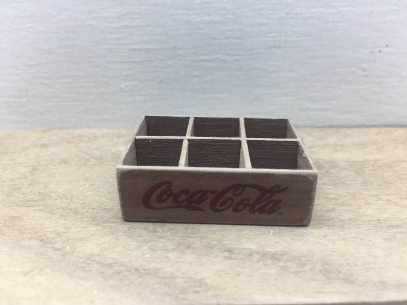 May include: A wooden Coca-Cola crate with six compartments. The crate is brown and has the Coca-Cola logo printed on the front in red.