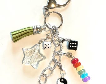May include: A silver keychain with multiple charms, including a clear glitter star, two dice, a yin-yang symbol, and a rainbow-colored bead strand. A green tassel hangs from the top. The keychain is designed to attach to a bag or set of keys.