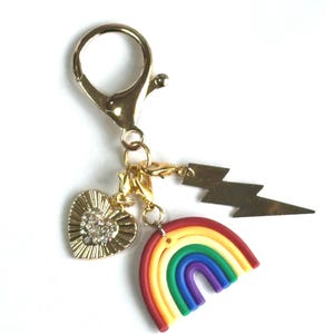 May include: Gold-toned keychain with a lobster clasp, featuring a heart charm with crystal accents, a rainbow charm, and a lightning bolt charm. The rainbow charm displays red, orange, yellow, green, blue, and purple colors.