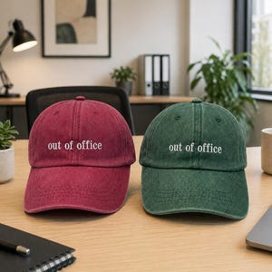 Könnte beinhalten: Zwei Baseballkappen, eine bordeauxfarbene und eine grüne, liegen nebeneinander auf einem Holzschreibtisch. Auf jeder Kappe steht in Weiß „out of office“. Die Kappen befinden sich vor einem Schreibtisch mit Büromaterial und einem Fenster.
