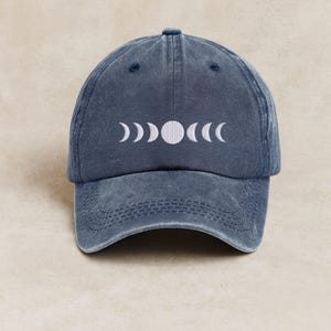 Op de afbeelding: Blauwe baseballcap met een wit geborduurd maanfase-ontwerp. De cap heeft een gebogen klep en een verstelbare band. Het maanfase-ontwerp bevat halve maan- en volle maanvormen.