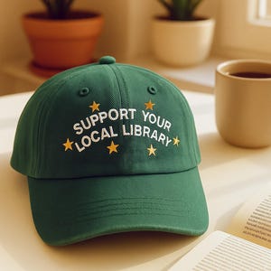 May include: A green baseball cap with the text "SUPPORT YOUR LOCAL LIBRARY" in white and gold stars. The cap has a curved brim and a button on top. The hat is sitting on a white surface.