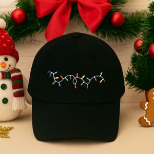 Könnte beinhalten: Schwarze Baseballkappe mit einer gestickten Lichterkette in verschiedenen Farben. Die Kappe wird mit einem Schneemann, einem Lebkuchenmann und Weihnachtsdekorationen präsentiert, was ein festliches Thema ergibt.