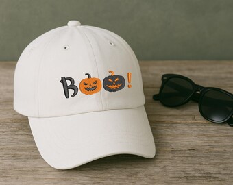 Op de afbeelding: Een witte baseballcap met het woord "Boo" in zwart geborduurd, met oranje en zwarte pompoenen. De cap heeft een gebogen klep en een knoop aan de bovenkant. Zwarte zonnebril ligt naast de cap.