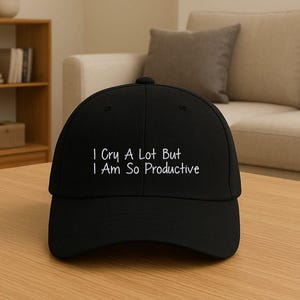 Könnte beinhalten: Schwarze Baseballkappe mit dem weißen Text "I Cry A Lot But I Am So Productive". Die Kappe liegt auf einem Holztisch. Im Hintergrund sind ein Sofa und ein Bücherregal zu sehen.